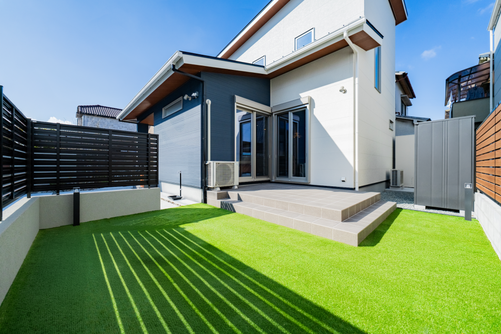 人工芝の庭とタイルテラスがある住宅の外観。掃き出し窓からつながるウッドフェンス付きプライベートガーデン