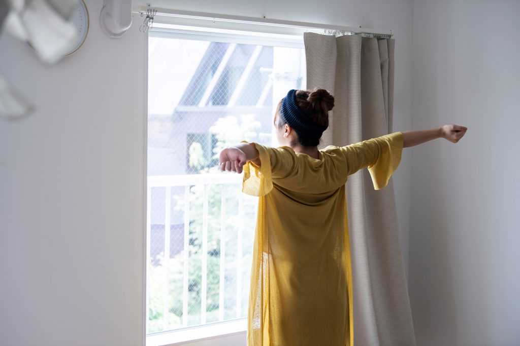 朝の光が差し込む窓の前で、両腕を広げて伸びをする女性の後ろ姿
